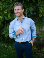 Man wearing a light blue shirt with rolled-up sleeves and navy chinos standing in front of green foliage.