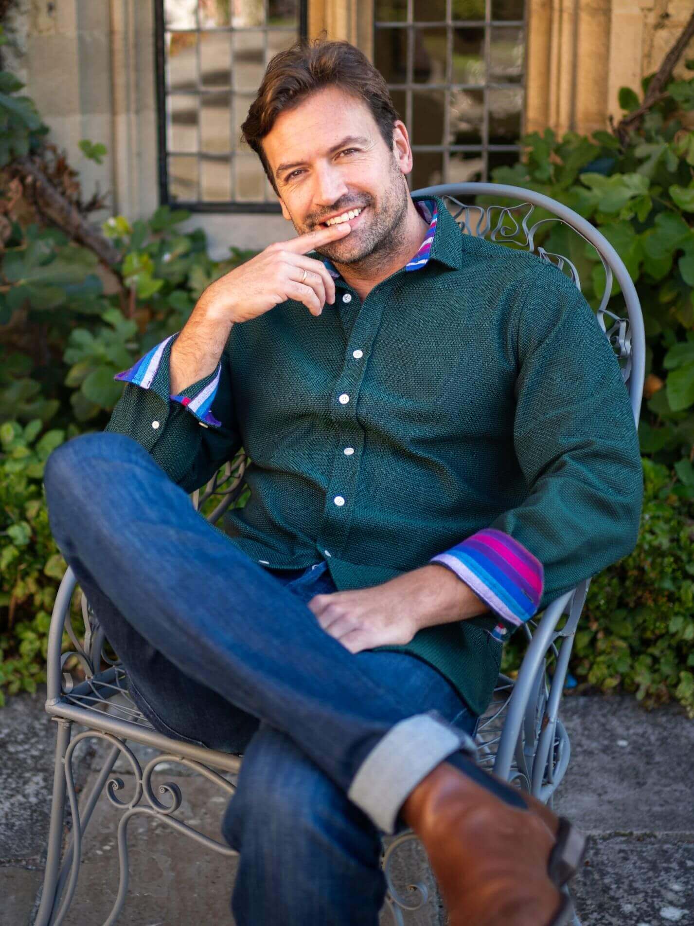 Man sitting outdoors in a garden, wearing a green basketweave shirt with colourful cuffs and blue jeans.