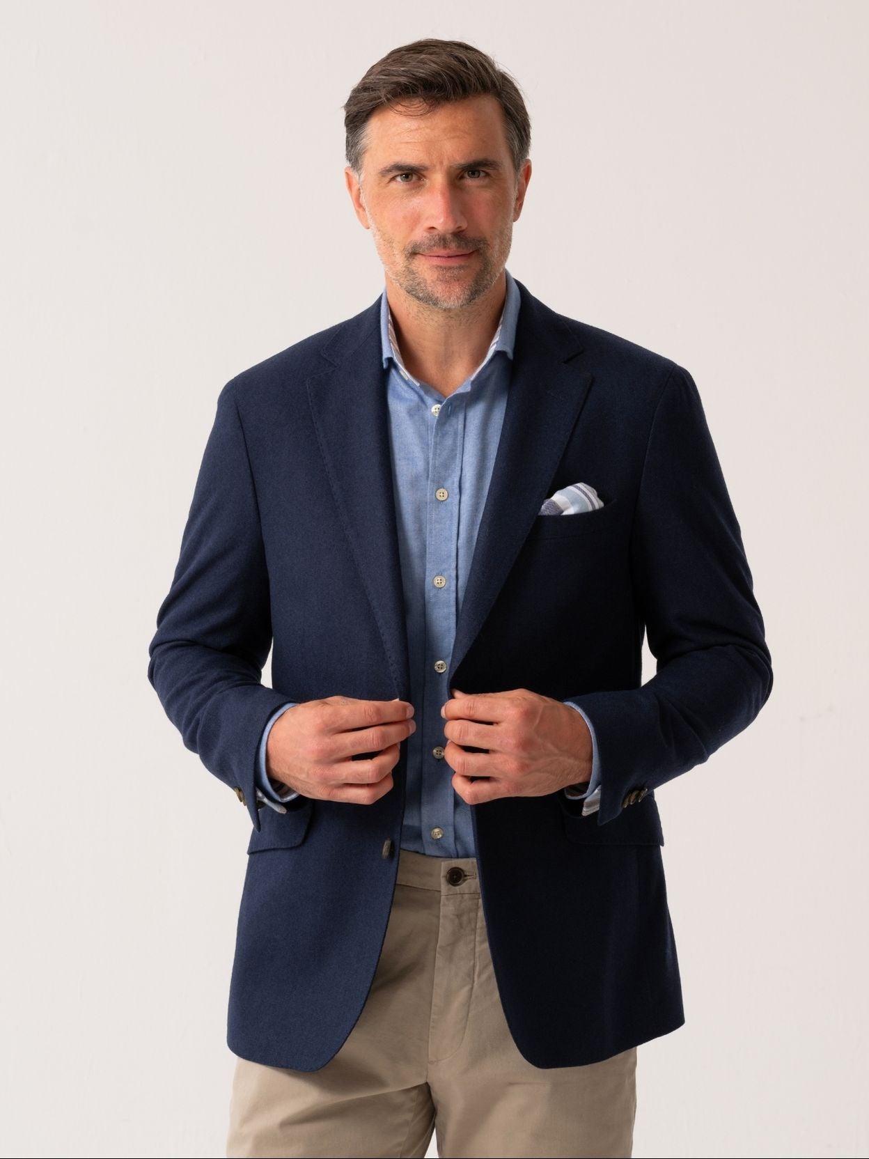 Man wearing a navy wool and cashmere blazer over a light blue shirt with beige trousers on a plain background