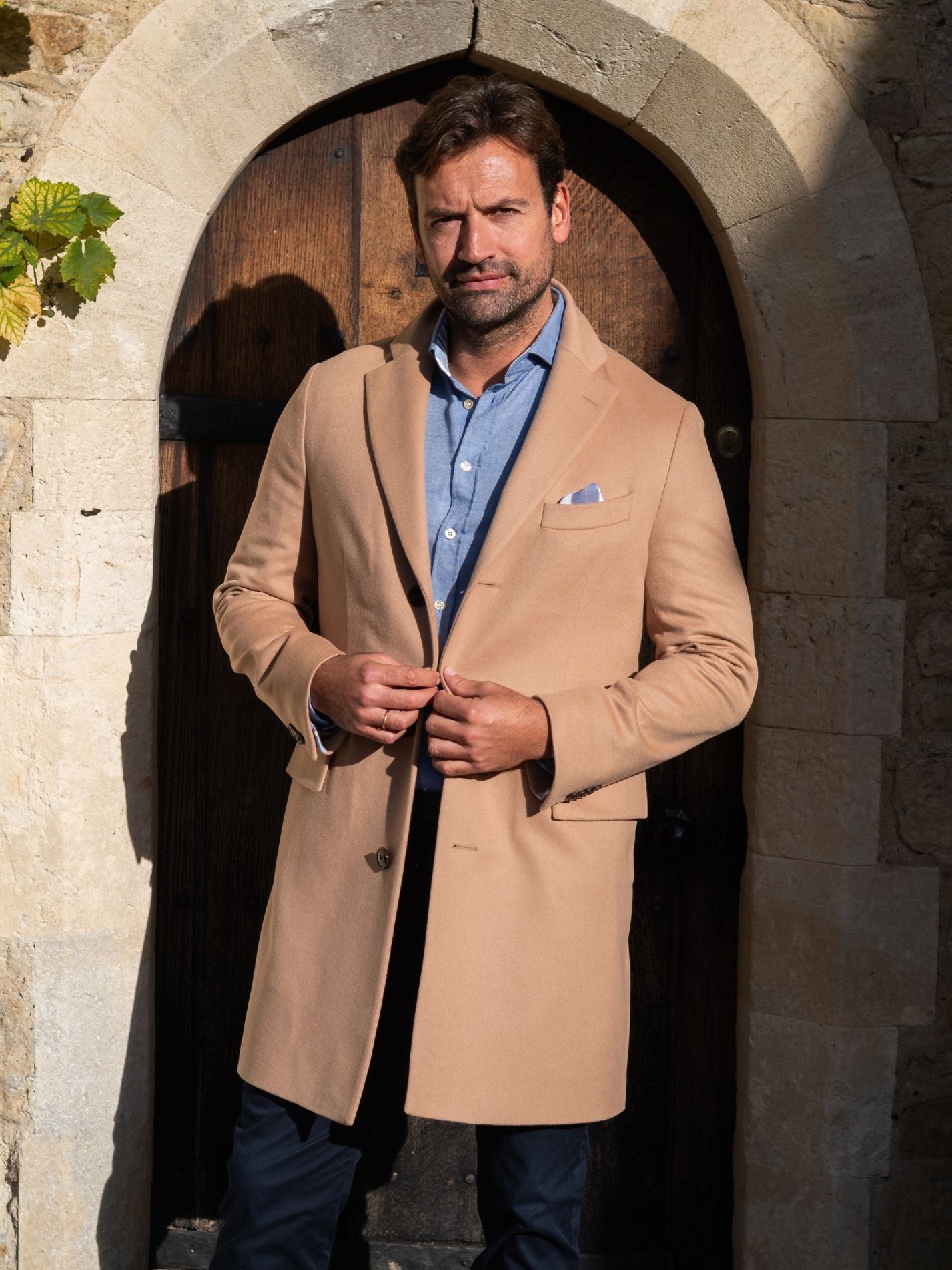 Man wearing a camel overcoat and blue shirt standing in front of a stone archway.