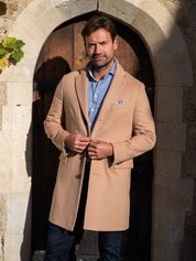 Man wearing a camel overcoat and blue shirt standing in front of a stone archway.