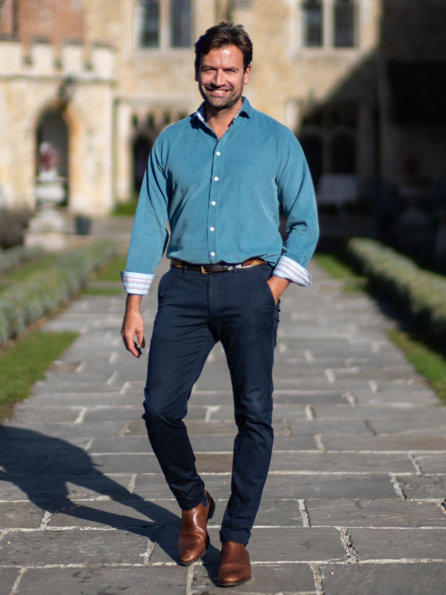 Man in a petrol blue shirt and navy chinos walking outdoors on a stone path.