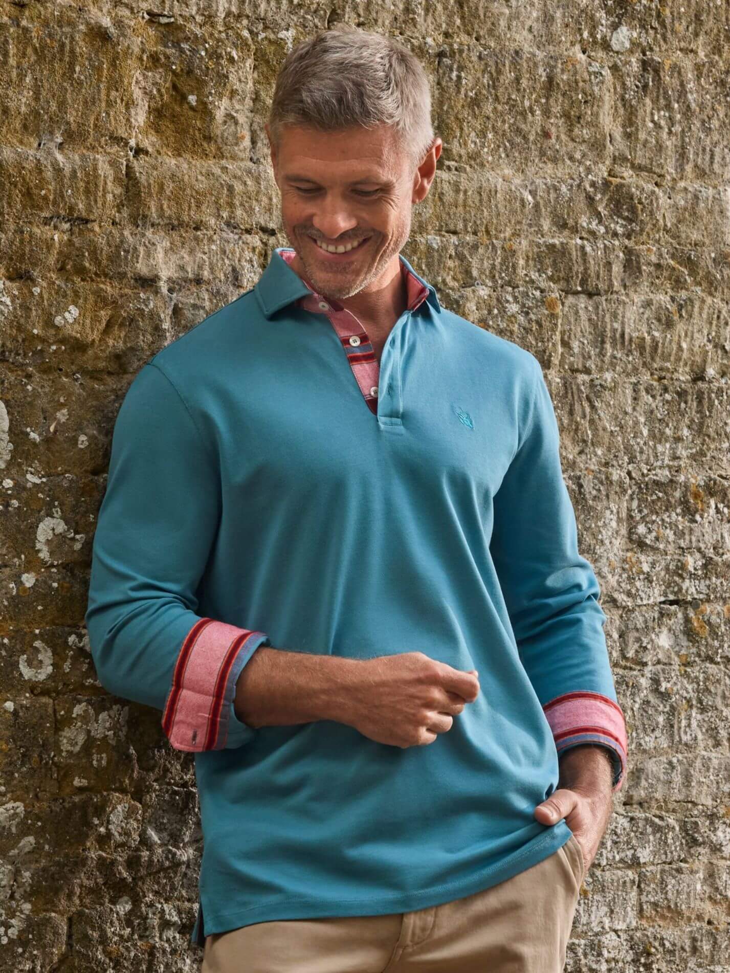 A man wearing a teal blue long-sleeve polo shirt with red and white striped cuffs, standing against a rustic stone wall.