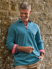 A man wearing a teal blue long-sleeve polo shirt with red and white striped cuffs, standing against a rustic stone wall.