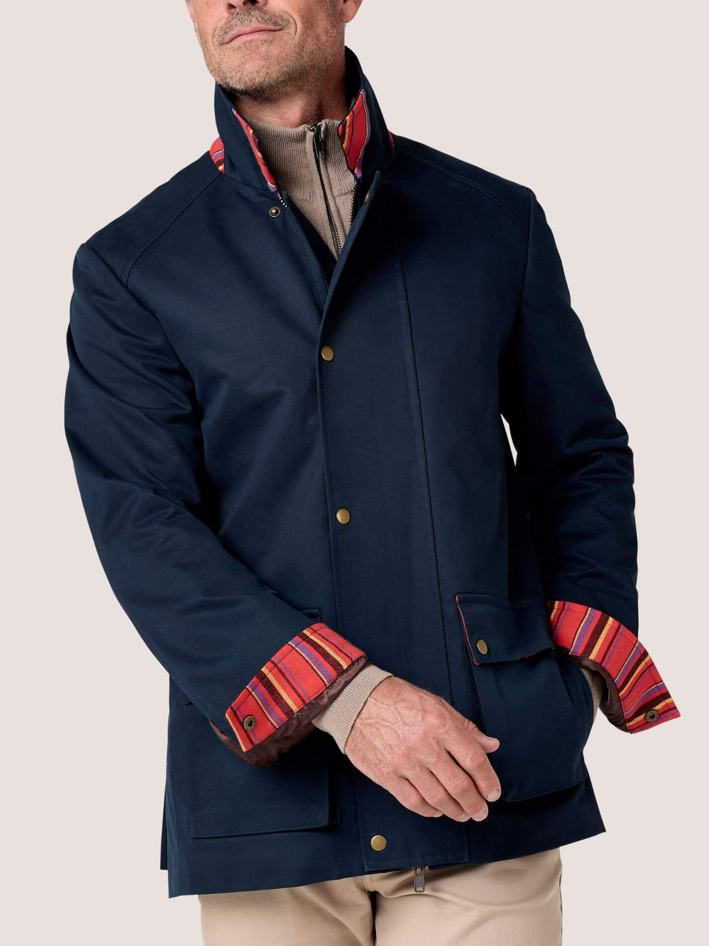A man wearing a navy blue jacket with a high stand collar and contrasting red, orange, and yellow striped inner lining visible at the collar and cuffs, paired with beige trousers against a light gray studio background.