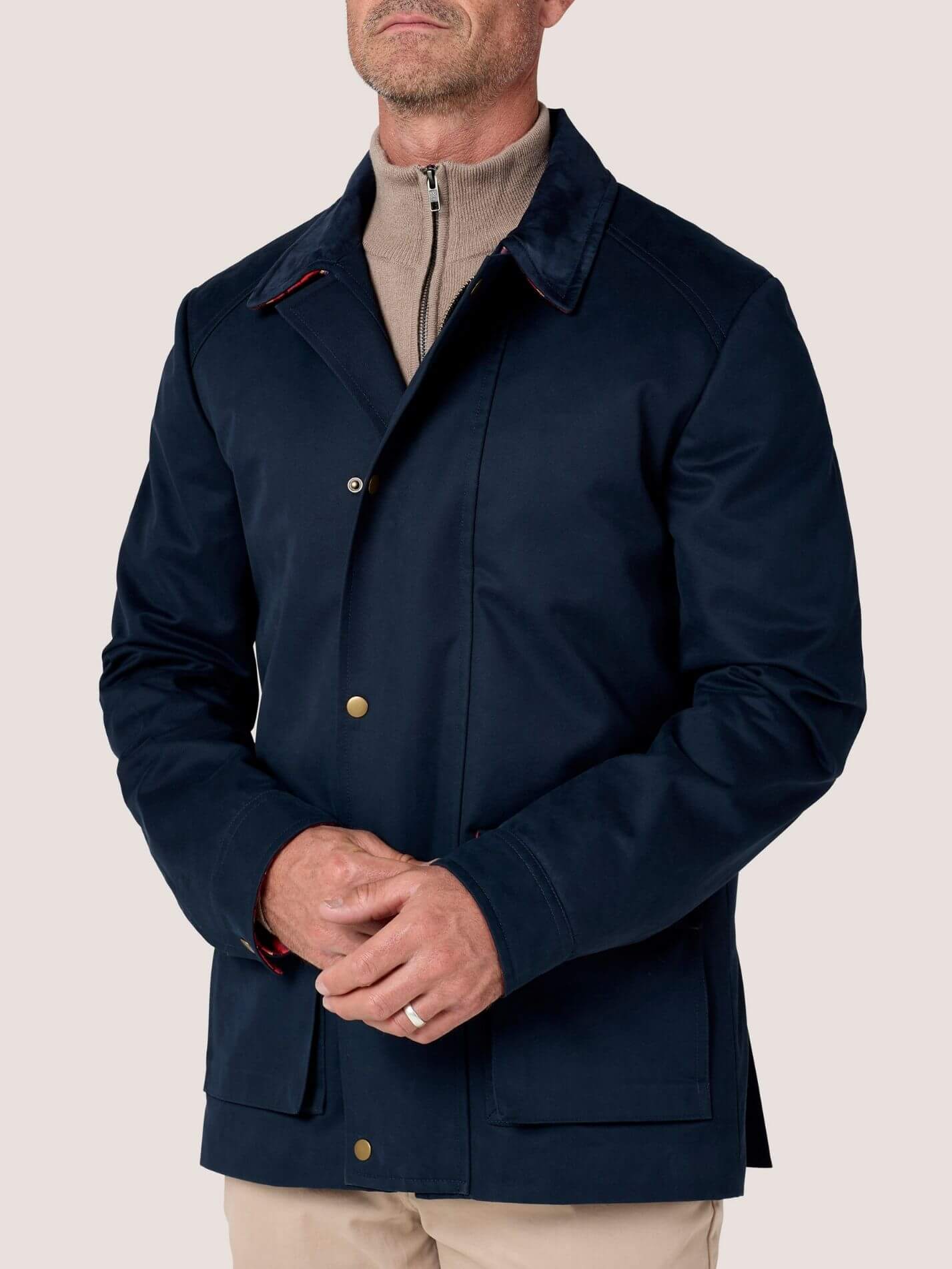 A man wearing a dark navy blue waxed canvas jacket over a beige half-zip sweater and beige trousers, shown from the chin down against a solid light beige background.