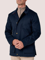 A man wearing a dark navy blue waxed canvas jacket over a beige half-zip sweater and beige trousers, shown from the chin down against a solid light beige background.