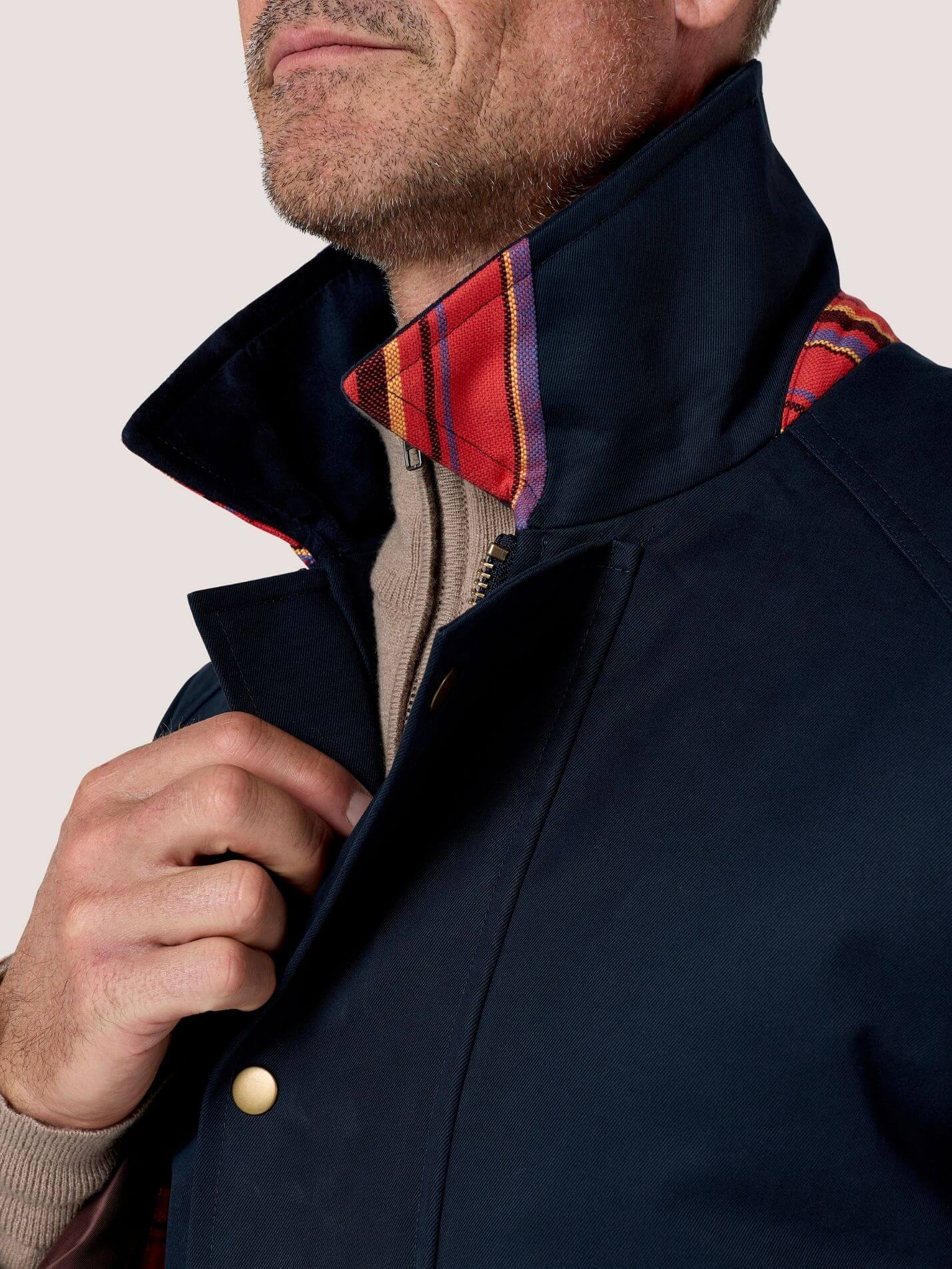 Close-up of a man wearing a navy blue jacket with a high collar, a red and purple plaid inner lining visible at the collar, and a beige knit shirt underneath.