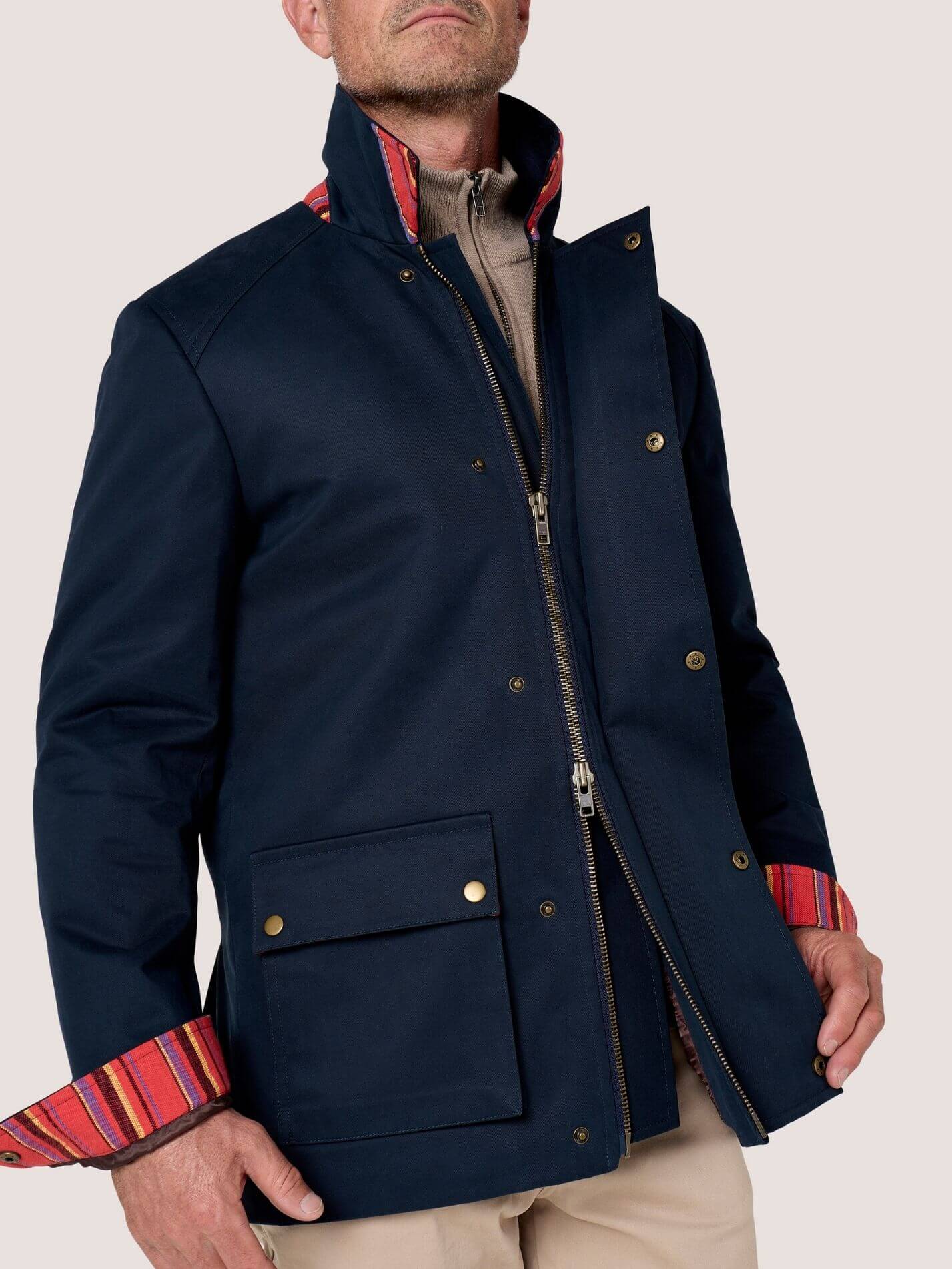A man wearing a navy blue jacket with a high collar, gold-tone snap and zipper details, and red striped knit cuffs, paired with beige trousers against a light beige background.