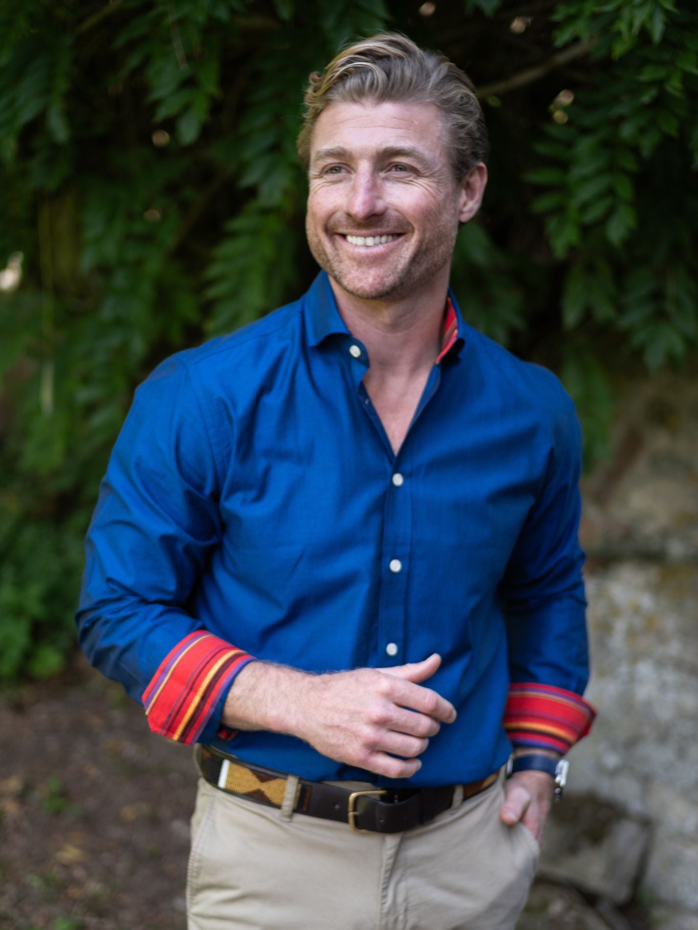 Man wearing a navy blue oxford cotton shirt with colourful cuffs standing outdoors against a green leafy background