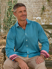 A man wearing a turquoise blue long-sleeve button-down shirt with pink striped cuffs, sitting on a wooden bench against a stone wall background.