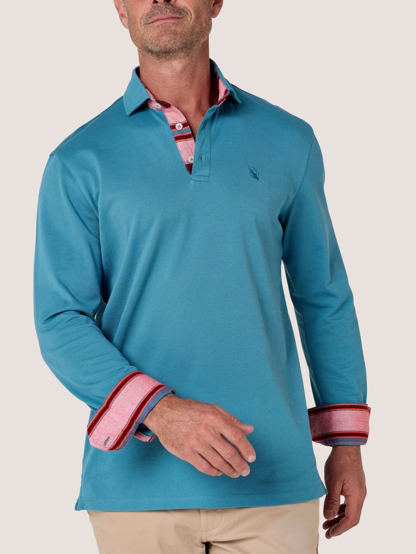 A man wearing a teal blue long-sleeve polo shirt with red and white striped cuffs, standing against a rustic stone wall.