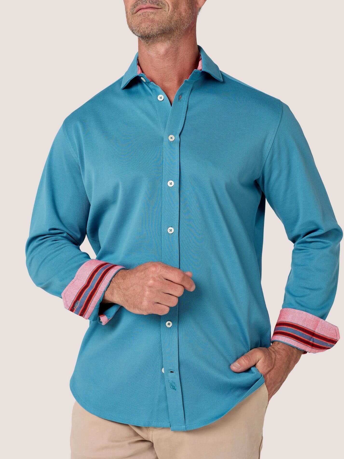 A man wearing a turquoise blue long-sleeve button-down shirt with pink striped cuffs, sitting on a wooden bench against a stone wall background.