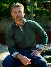 A man wearing a dark green long-sleeve polo shirt over a striped collared undershirt, sitting outdoors in a garden setting.